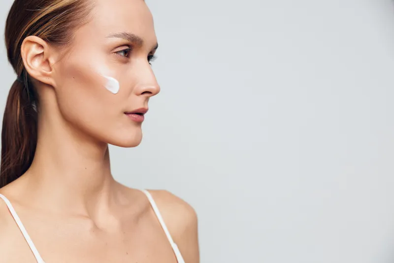 A young woman with clear skin applies white cream on her cheek. She looks to the side against a plain light background. Concept for skincare, beauty, and facial care.