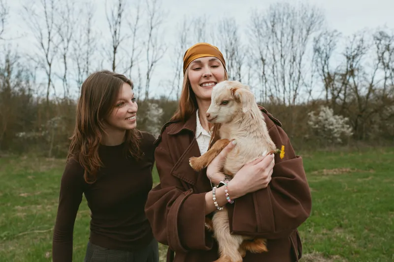 Cheerful outdoor lifestyle moment with two women smiling, one cradling a playful goat kid, warm coat and hat, soft natural light, relaxed countryside setting, candid friendship and gentle animals