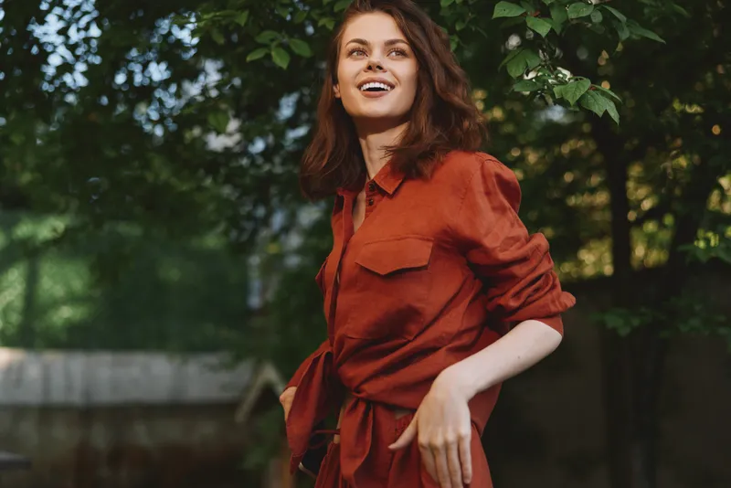 Smiling woman in a red shirt outdoors, casual lifestyle portrait among green trees, warm sunlight, candid moment, fresh air, relaxed mood for lifestyle and outdoor living vibes