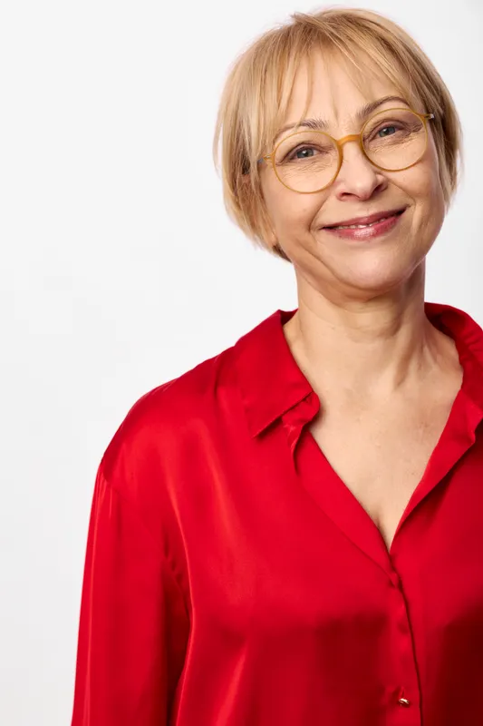 Senior woman portrait, smiling with glasses, short blonde hair, wearing red blouse against white background, friendly mood, studio headshot, soft light.