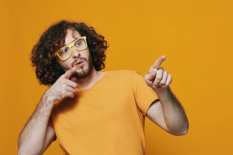 Young man with curly hair in yellow glasses pointing at the camera against a vibrant orange background, expressing curiosity and creativity in a playful manner