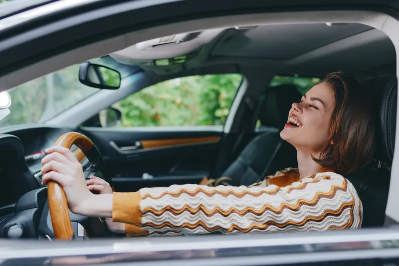 A joyful woman drives a car, smiling as she grips the wooden steering wheel, enjoying a candid moment inside a stylish cabin.