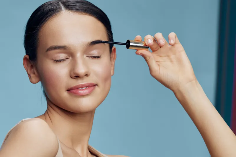Young woman applying mascara with a joyful expression on a pastel blue background, showcasing beauty and confidence in cosmetics
