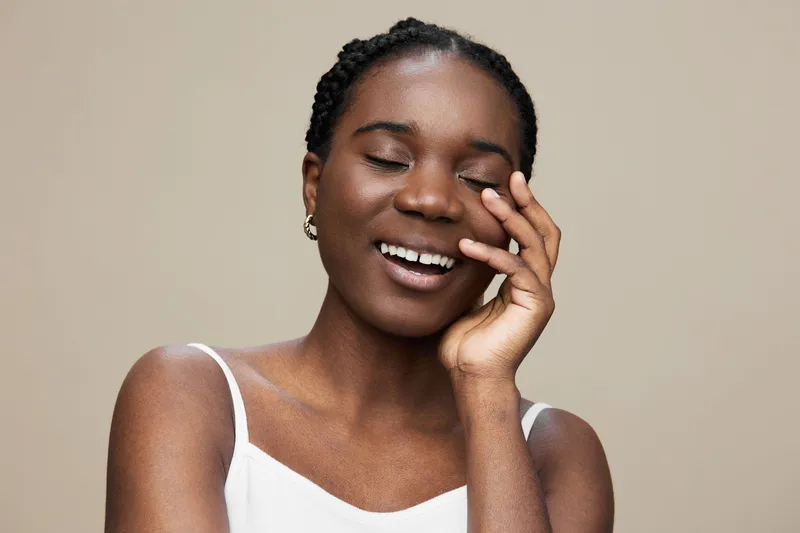 Joyful young Black woman smiling with closed eyes, wearing a white top, against a soft beige background, representing happiness and positivity