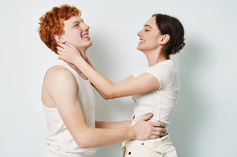 Young couple with happy expressions embracing and smiling isolated on white background. Woman holding man's neck and man hugging woman in casual white outfits.