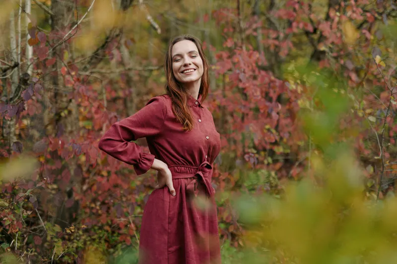A smiling woman in a simple red dress stands among autumn foliage, conveying a natural, unposed presence and authenticity that emphasizes genuine emotion and everyday beauty in a quiet outdoor
