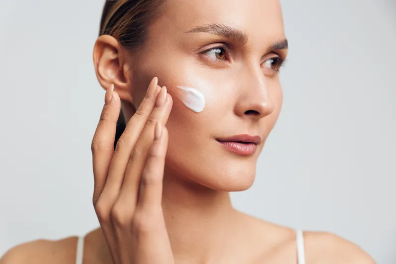 Young woman applying white face cream on cheek with fingers, close-up portrait on light background. Concept of skincare, beauty routine, moisturizing, and healthy skin care.