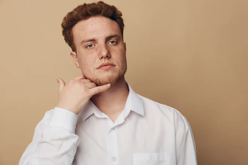 Young man in white shirt makes call me gesture with hand near face, looking at camera on beige background. Concept of communication, gesture, and casual style portrait.