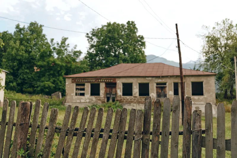 Rural house with weathered walls sits behind a leaning wooden fence in a quiet countryside setting, conveying forgotten charm and a rustic tranquil atmosphere under a cloudy sky
