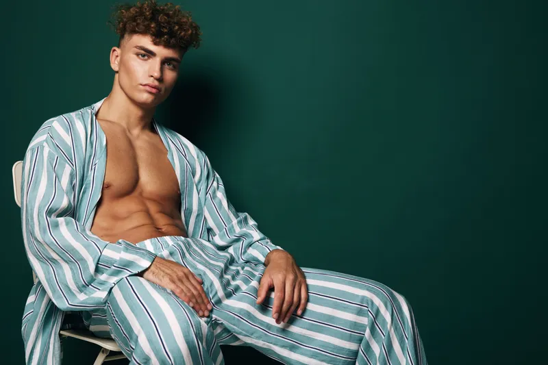 Young man with curly hair wearing striped pajamas sitting on chair against dark green background. Casual pose, relaxed mood, and fashion style with bare chest and comfortable outfit.