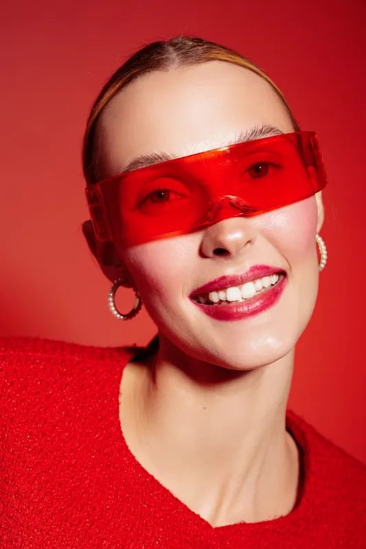 Smiling woman with red sunglasses and red sweater poses against red background. Close-up portrait with hoop earrings and glossy lips. Concept of fashion, style, and confidence.