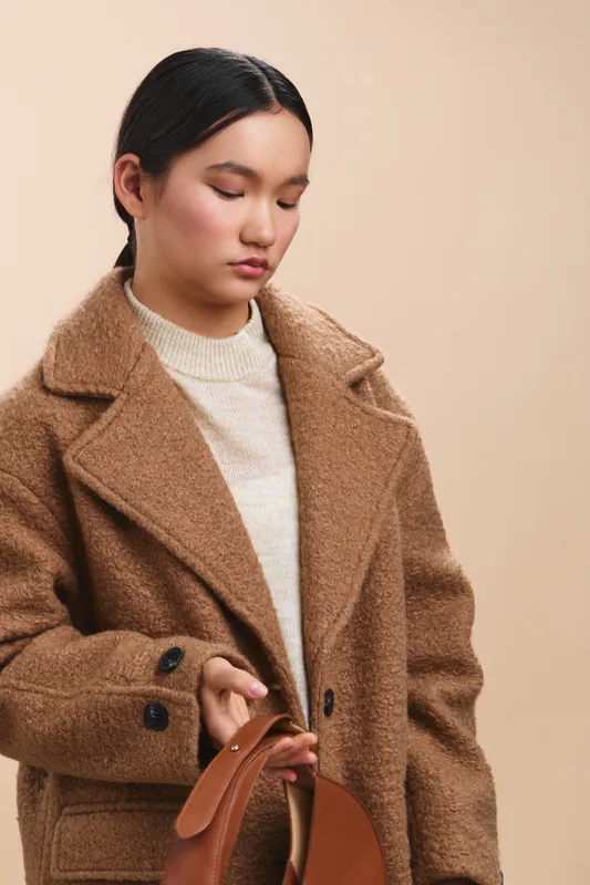 asian woman wearing a brown coat stands against a neutral background, looking down at a handbag with a calm expression, conveying a stylish, minimalist lifestyle moment.