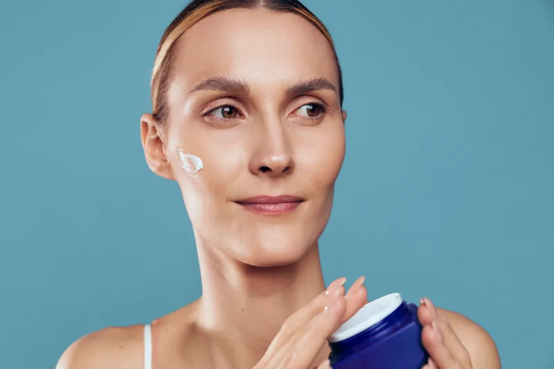 A woman with clear skin applies cream on her cheek while holding a blue jar. The close-up shot shows skincare routine and facial care with a calm expression on a blue background.