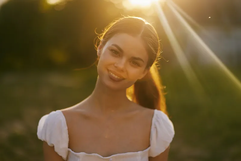 Beautiful young woman with long hair smiling in a sunny field during golden hour, wearing a white dress, evoking joy and warmth in a natural setting