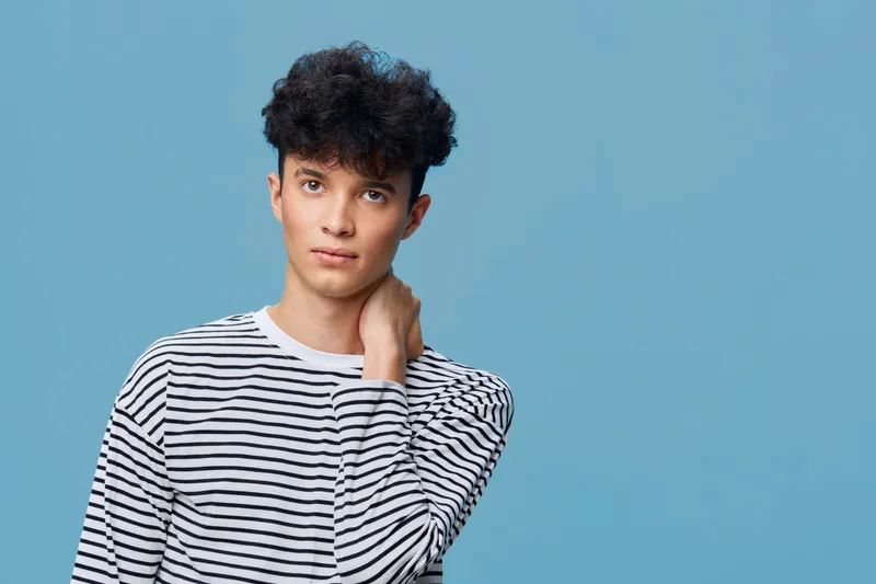 young man in striped shirt looking thoughtfully, casual style with curly hair and blue background, portrait showing natural expression and relaxed pose