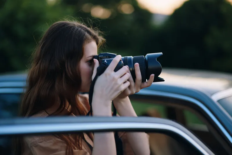 A young woman capturing photos with a professional camera, emphasizing creativity and passion in photography during golden hour near a classic car.