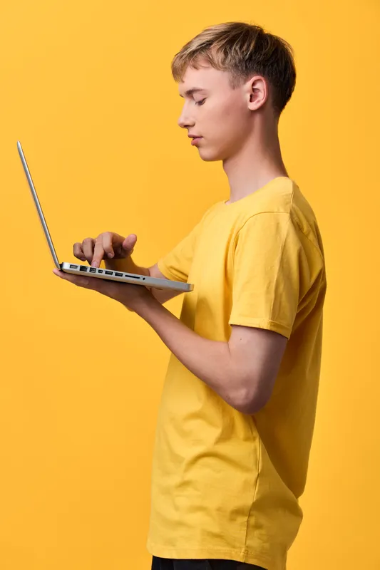 Young boy in a yellow tee types on a slim laptop against a vibrant background, exploring technology and online learning with focus on screen, creativity, and digital skills