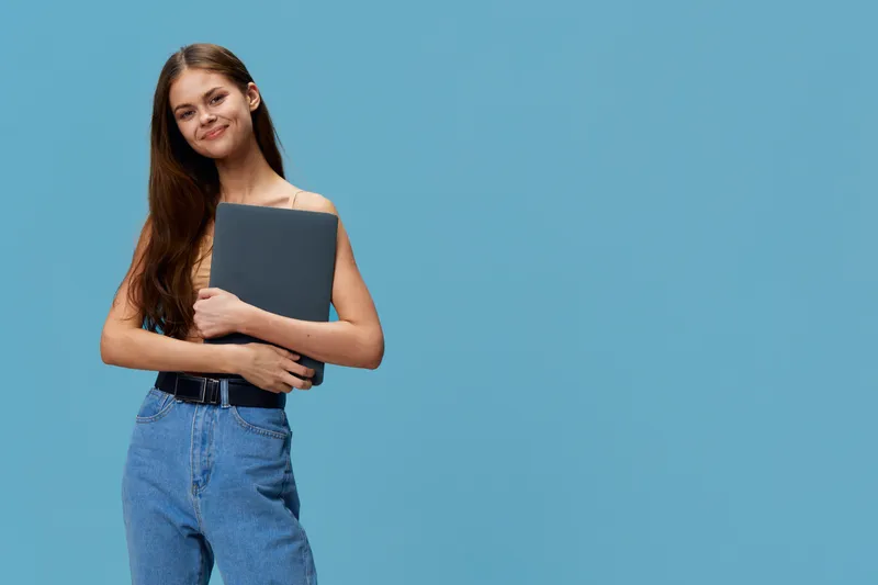 smiling young woman holding laptop wearing casual clothes standing against blue background, showing relaxed confident pose and natural long hair