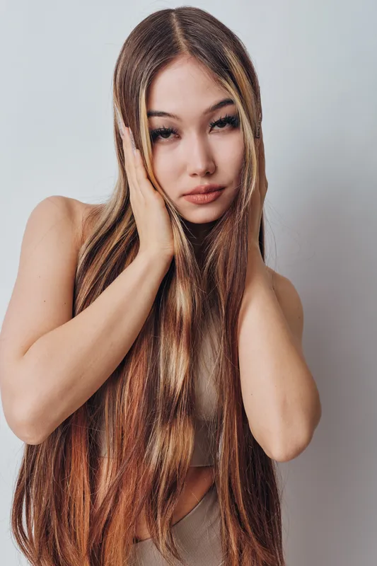 Young woman with long straight hair posing with hands on face, natural expression and smooth skin, studio portrait on neutral background.