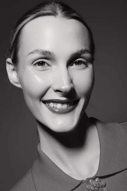 Close-up portrait of a smiling Caucasian woman with fair skin and smooth hair, wearing a collared top. Studio lighting highlights her natural beauty and confident expression.