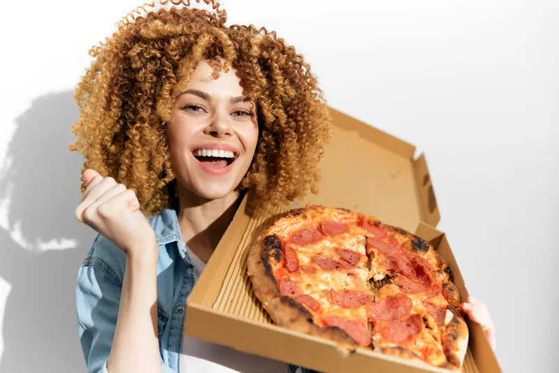 Радостная молодая женщина с кудрявыми волосами наслаждается вкусной пиццей