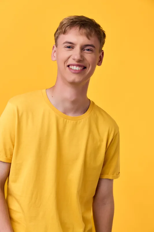 Bright studio shot of a young man in a yellow tee, standing against a vivid yellow backdrop with a friendly smile and casual pose, conveying positivity and energy.