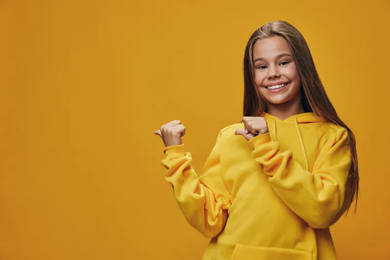 Girl smiling and wearing yellow hoodie with long hair standing against yellow background pointing at herself with both thumbs. Happy child with confident expression and casual style.