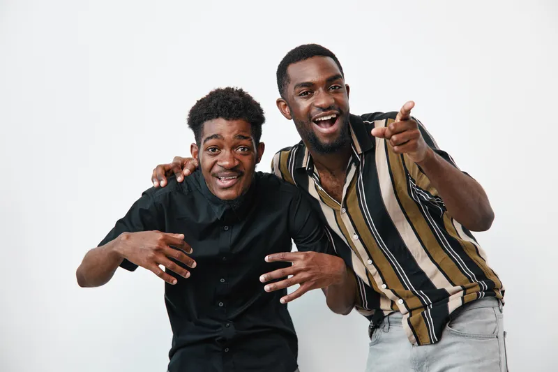 Two young black men with dark skin posing together on white background smiling and laughing. Casual clothing and friendly gesture showing happiness and friendship.