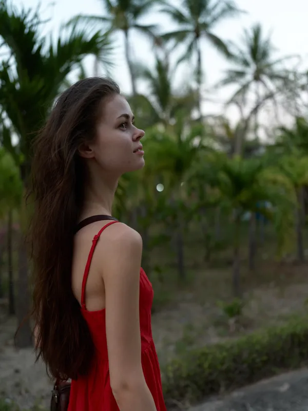 young woman in red dress standing outdoors among palm trees, looking thoughtfully into the distance, natural light highlighting her profile and long hair