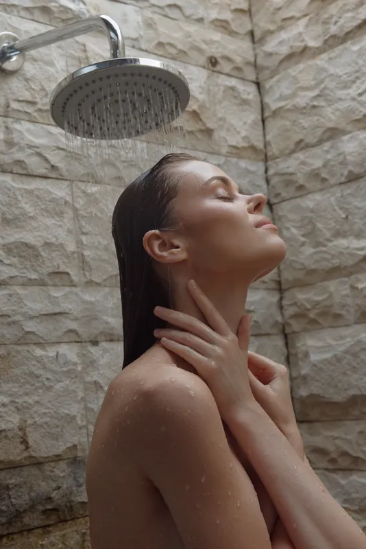 young woman enjoying refreshing outdoor shower with natural stone wall background, wet hair, relaxed expression, wellness and skincare moment