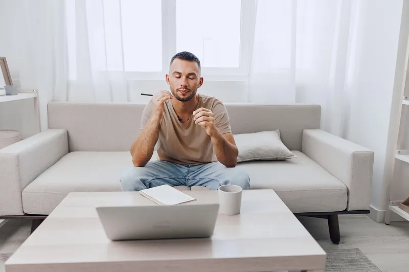 Focused man in casual attire, sitting on a sofa with a laptop, jotting down ideas in a notebook, surrounded by a calm and cozy workspace atmosphere.