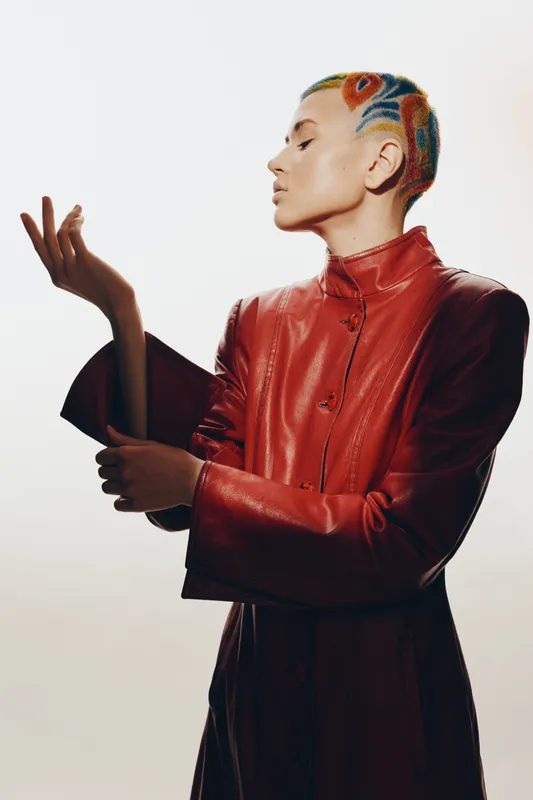 Woman with colorful patterned shaved hair wearing a red leather coat, posing elegantly against a plain white background, showcasing bold fashion and unique style.