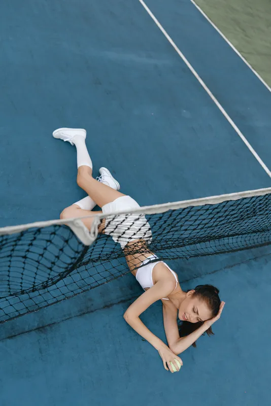 Young female tennis player lying on the court, showcasing exhaustion and determination, sporting athletic wear, with a vibrant blue background and seasonal outdoor setting