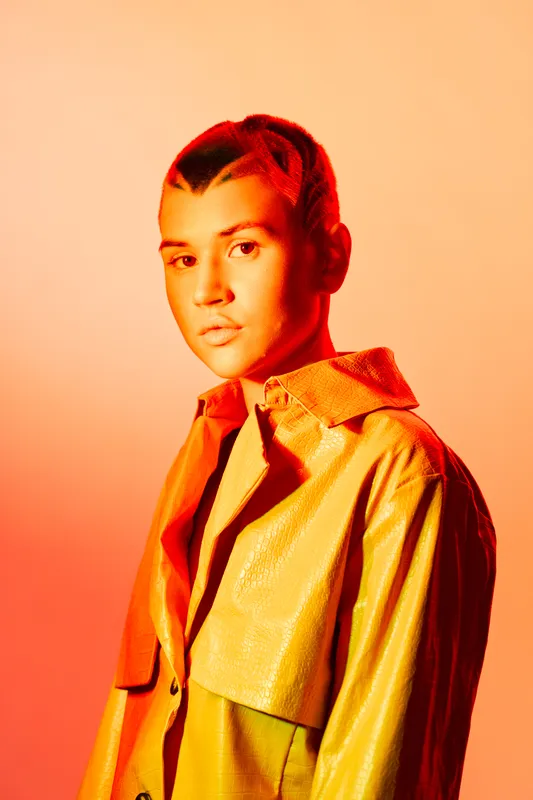 young woman with short hair wearing a yellow jacket and posing with a serious expression against an orange gradient background in studio lighting