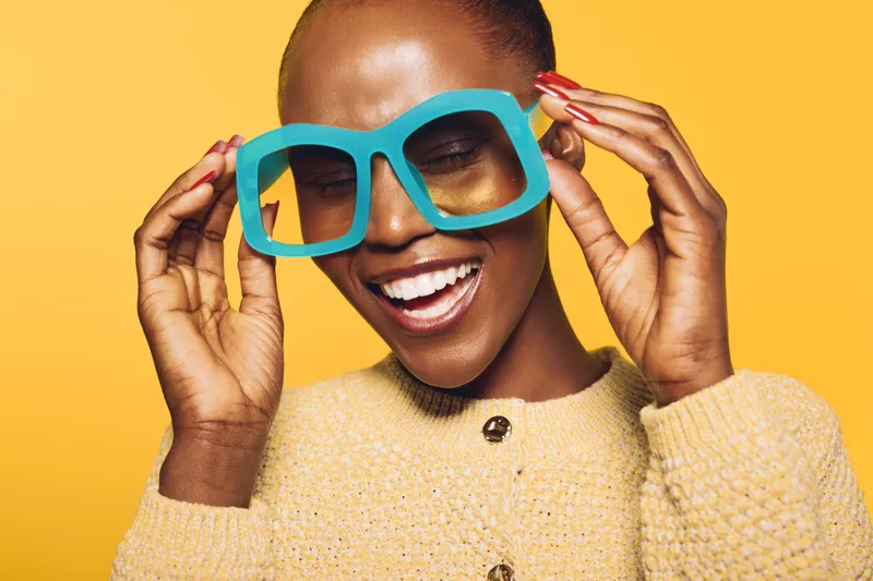 Happy African woman with dark skin smiles broadly while holding large blue glasses on yellow background. Concept of joy, fashion, and vibrant lifestyle in studio portrait.