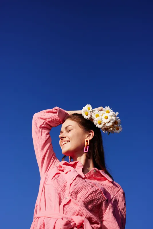 Cheerful young woman with floral headband smiling in bright pink outfit against a clear blue sky, exuding happiness and spring vibes