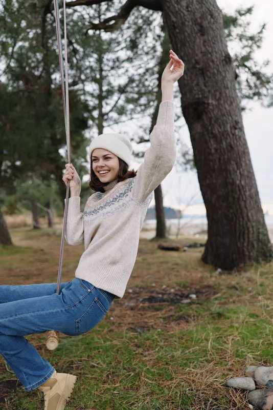 Woman on a rope swing outdoors in a park, smiling and enjoying a playful moment in a cozy sweater and beanie, surrounded by trees and grassy ground.