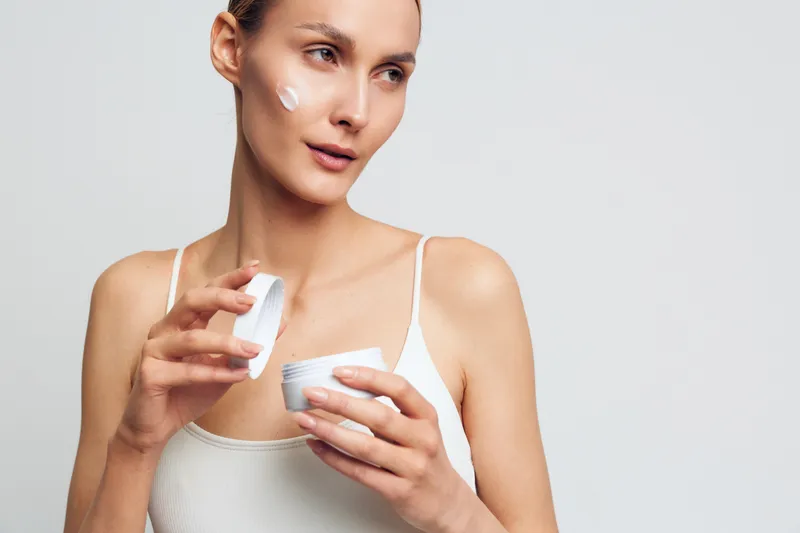 Young woman applying face cream with soft texture on cheek, skincare routine, natural makeup, white tank top, clean background, focused expression, beauty care concept