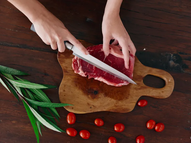 Женщина с ножом в руке нарезает свежее стейковое мясо для жарки на кухне