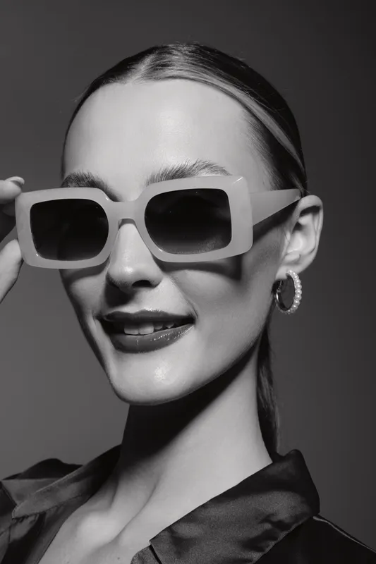 Portrait of a young white woman with fair skin wearing large sunglasses and hoop earrings smiling confidently against a plain background studio fashion concept.
