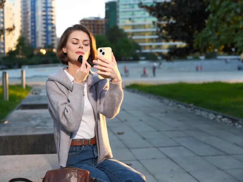 A stylish young woman sits in a modern urban park, applying lipstick as she checks her phone, smiling softly and enjoying a casual outdoor moment.