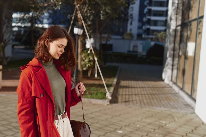 A woman in a red coat walks outdoors, head bowed, adjusting her strap as sunlight washes the modern plaza, creating a candid urban fashion moment.