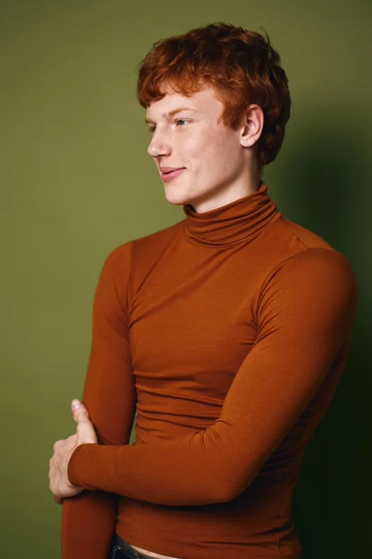 Young man with red hair wearing orange turtleneck posing with arms crossed, looking sideways on green background for fashion portrait and style.