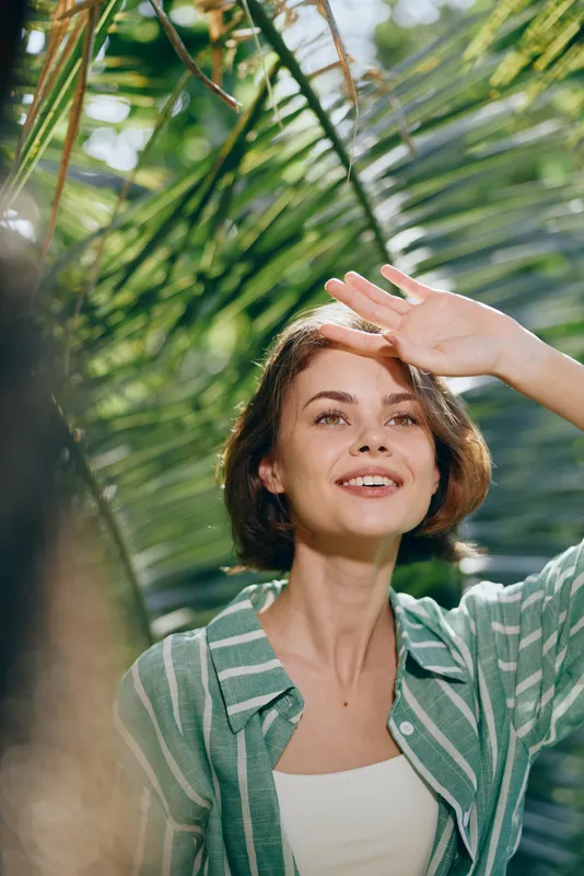 Woman portrait outdoors under bright sunlight, shielding her eyes with a raised hand, wearing a striped green shirt, surrounded by lush greenery and tropical leaves.