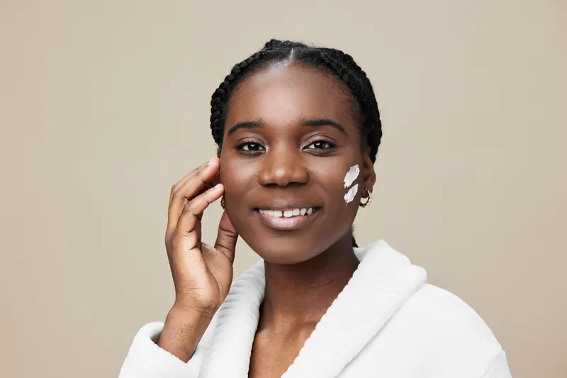 Smiling Black woman applying skincare cream on her face, dressed in a cozy white robe, against a neutral beige background emphasizing skincare and beauty