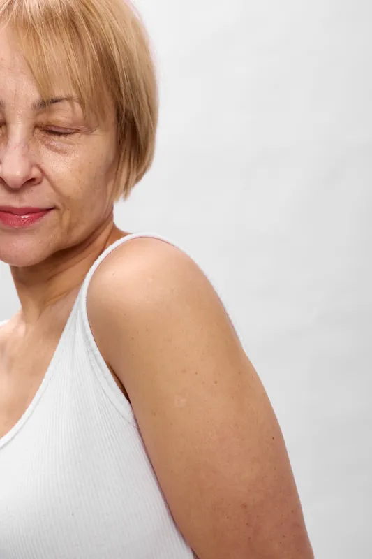 Mature woman portrait closeup, eyes closed, wearing sleeveless white top, calm expression, hand at shoulder, soft studio light, plain white background, relaxed