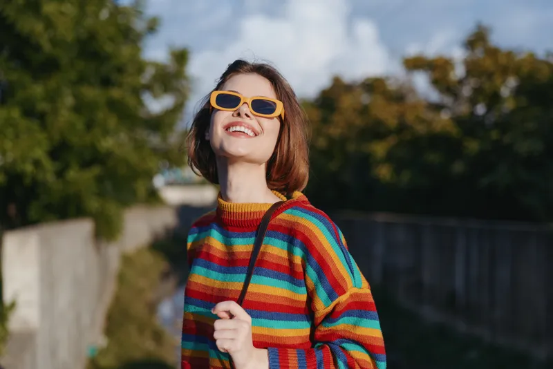 Woman smiling in a colorful striped sweater and orange sunglasses outdoors, young adult with short hair looking up, casual outfit and joyful expression in urban nature for lifestyle travel.