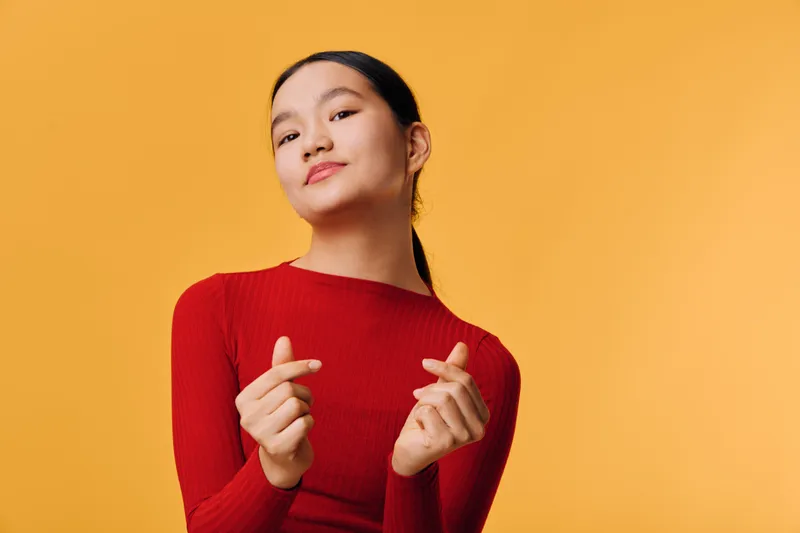 Young woman in red sweater making finger heart gesture smiling confidently with clean yellow background studio portrait for positive emotions and modern fashion style.