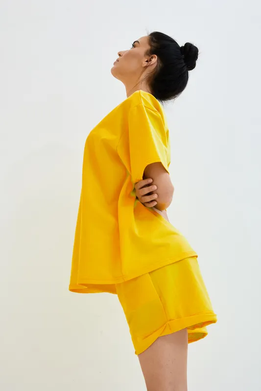Young woman wearing bright yellow casual t-shirt and shorts posing in profile with arms behind back on plain white background, showing relaxed and confident posture.