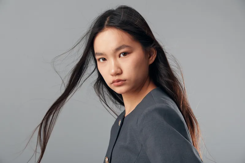Young Asian woman posing with serious expression looking at camera with hair flowing and wearing dark jacket on plain grey background in studio lighting.